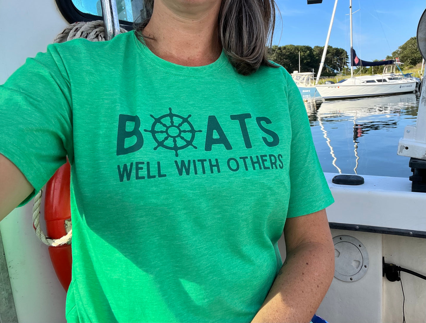 Person wearing the Boats Well With Others unisex tee in kelly green with blue graphic, enjoying a day on the water aboard a boat