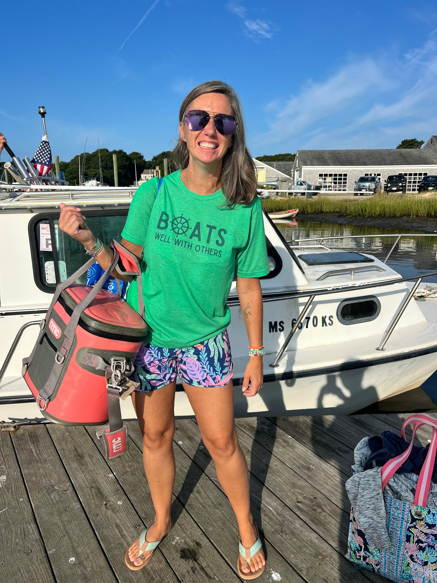 Model standing in front of a boat wearing the Boats Well With Others unisex tee in kelly green with blue nautical graphic