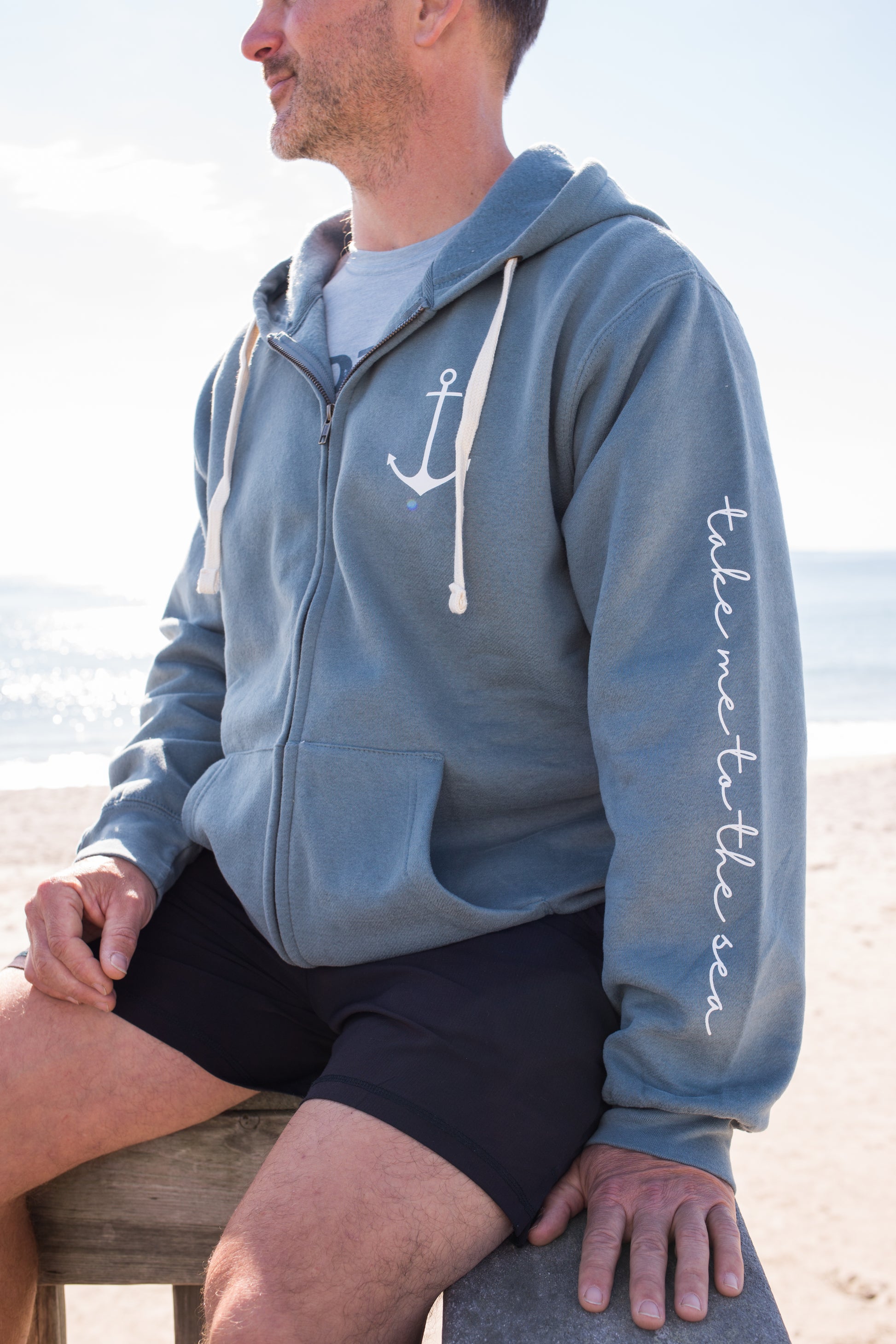 Model wearing marine blue heavyweight zip hoodie with anchor chest logo and 'take me to the sea' quote on sleeve, showcasing relaxed unisex fit.